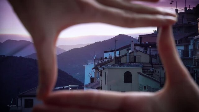 Finger Frame. Woman folding her hands in frame with a setting sun illuminates the roofs of a medieval town in the mountains. Perinaldo, Italy.