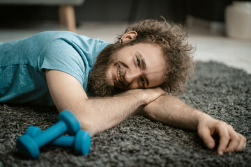 Man Lies Relaxed on the Carpet Next to his Dumbbells. Curly-haired Guy is too Relaxed to Play Sports. Close-up Portrait  - obrazy, fototapety, plakaty