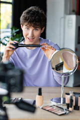 Smiling transgender makeup artist holding cosmetic brush near cosmetics and blurred digital camera.