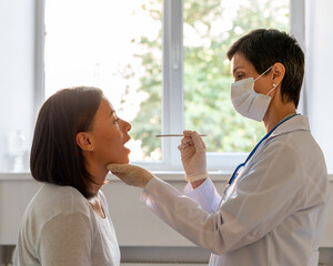 Middle-aged doctor in protective mask examining patient throat and oral cavity