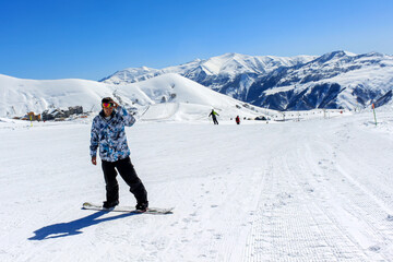 A man snowboarding on a ski slope. In the background is a blue sky and snowy mountain tops. Active...