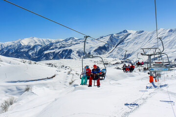 Fototapeta na wymiar People on a chairlift ascend a ski slope. In the background snowy mountain ridge and clear blue sky. Active winter holidays in the mountains.