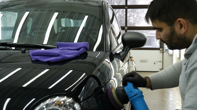 A man in a special suit polishes a gray car body, a tool for polishing cars, into a workshop.	
