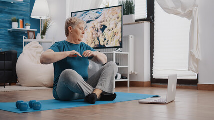 Fototapeta na wymiar Senior woman following training video program on laptop to do workout and physical activity. Retired person watching online lesson with trainer to exercise fitness and gymnastics on mat.