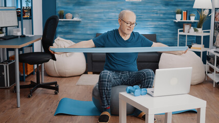 Aged man pulling resistance band to stretch arms muscles and following training lesson on laptop while sitting on fitness toning ball. Old person using elastic belt and workout video.