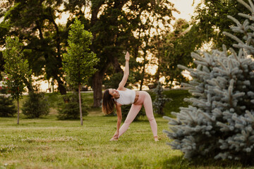 attractive, elegant woman do yoga and stands in Trikonasana pose while maintaining balance on green grass of lawn at sunset, concept of inner harmony. 