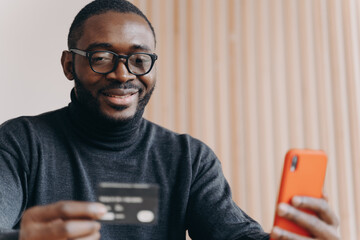 Young positive African ethnicity man entrepreneur in glasses paying with credit card online