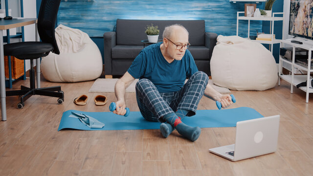 Senior adult using dumbbells to do physical exercise while watching workout video on laptop. Aged man following online training lesson on computer and lifting weights to do fitness activity.