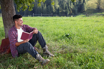 Naklejka na ściany i meble Handsome man reading book under tree on green meadow near forest