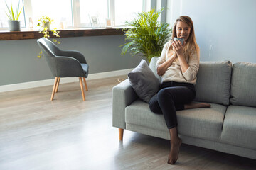 Young coffee lover sniffing hot drink at cozy home interior. Woman relaxing on the sofa at home with a cup of coffee. Coffee and a quiet moment. Shot of a beautiful young woman having a cup of coffee