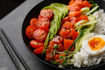 Bright Salmon Poke bowl with avocado, cherry, egg and rice on dark background with chopsticks. Top view and close up
