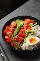 Close-up Salmon Poke bowl with avocado, cherry, egg and rice on dark background with chopsticks.