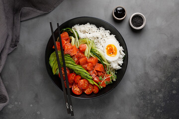 Salmon Poke bowl with avocado, cherry, egg and rice on dark background with chopsticks. Top view
