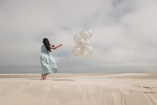 Girl in light blue dress holding bunch of a white balloons om windy day on beach dune