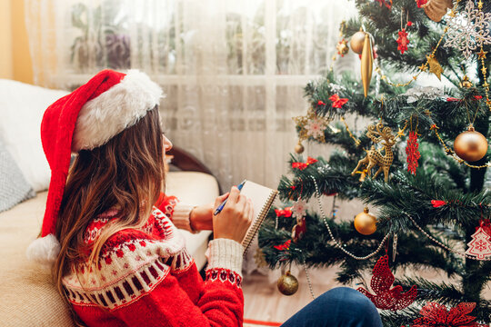 Young woman writing to do list for 2022 New year in notebook at home sitting by Christmas tree in Santa hat.