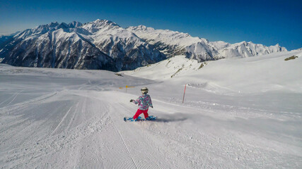 A snowboarder going down the slope in Heiligenblut, Austria. Perfectly groomed slopes. High mountains surrounding the girl, wearing pink trousers and colorful jacket. Girl wears helm for protection