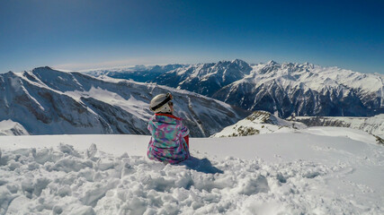A snowboarder girl taking a break on the slope. The snowboard is white and blue, harmoniously fitting to the nature conditions, white snow, blue sky. There are endless chains of mountains in the back