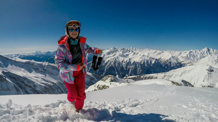 A girl in a snowboarding outfit and a helm walks on a thick snow. She is smiling. Thick layer of snow covering the mountains lopes. Tall mountains in the back. Girl is enjoying the view and the day.