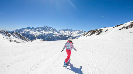 Fototapeta na wymiar A snowboarder going down the slope in Heiligenblut, Austria. Perfectly groomed slopes. High mountains surrounding the girl, wearing pink trousers and colorful jacket. Girl wears helm for protection