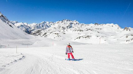 A snowboarder going down the slope in Heiligenblut, Austria. Perfectly groomed slopes. High mountains surrounding the girl, wearing pink trousers and colorful jacket. Girl wears helm for protection