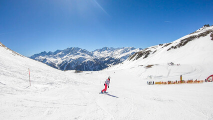 A snowboarder going down the slope in Heiligenblut, Austria. Perfectly groomed slopes. High mountains surrounding the girl, wearing pink trousers and colorful jacket. Girl wears helm for protection