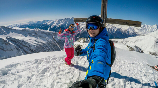 A couple in skiing outfit stands under the cross on top of a snowy mountains. Man is holding a selfie stick and takes the picture. Tall Alp's peaks behind the couple. Happiness and fun while skiing.