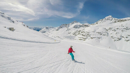 A snowboarder going down the slope in Moelltaler Gletscher, Austria. Perfectly groomed slopes. High mountains surrounding the girl wearing colourful snowboard outfit. Girl wears helm