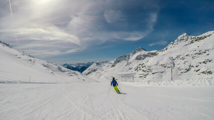 A skier going down the slope in Moelltaler Gletscher, Austria. Perfectly groomed slopes. High mountains surrounding the man wearing yellow trousers and blue jacket. Man wears helm for the protection.