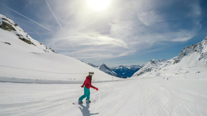 A snowboarder going down the slope in Moelltaler Gletscher, Austria. Perfectly groomed slopes. High mountains surrounding the girl wearing colourful snowboard outfit. Girl wears helmet