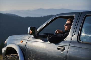 Against the backdrop of forest and mountain hills in distance, view of SUV driver leaning with one hand on side door examining beautiful scenery in sunglases while driving off-road.
