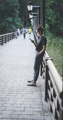 Candid side view point of young woman standing on the bridge in public park and playing with her phone , hope and dream concept