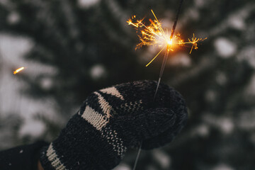 Happy New Year! Hand in cozy glove holding burning sparkler on background of pine tree branches in snow. Atmospheric magic moment. Woman hand with glowing firework in evening. Happy Holidays!
