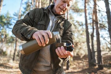 Old man pouring hot beverage to the cup from the thermos while resting