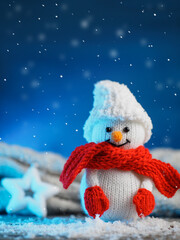 Knitted toy snowman in a hat, mittens and scarf. Blue background with falling snow