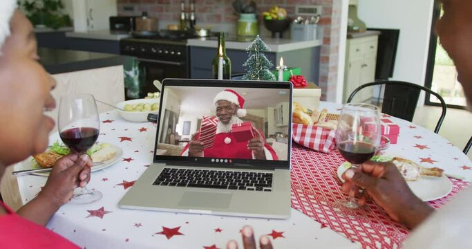 African american couple with wine using laptop for christmas video call with happy santa on screen