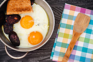 Sartén con dos huevos fritos, morcilla y una rebanada de pan, servilleta de colores y espatula de cocina, visto desde arriba
