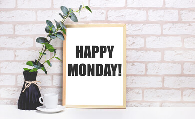 On a white table against a light brick wall, a branch of eucalyptus in a dark vase, a white cup and a light wooden frame with the text HAPPY MONDAY. Home office interior.
