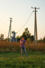 Epic portrait of young brunette man in purple polo shirt standing outdoor on nature background at summer day. Millennial generation. Outside. Overall plan. Electric pole and transformation
