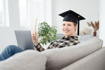 Happy woman holding diploma scroll, wearing black academic cap on her head, at home, laptop on her...