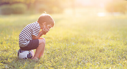 A little boy sits and is sad on a grassy field with a sun glare.