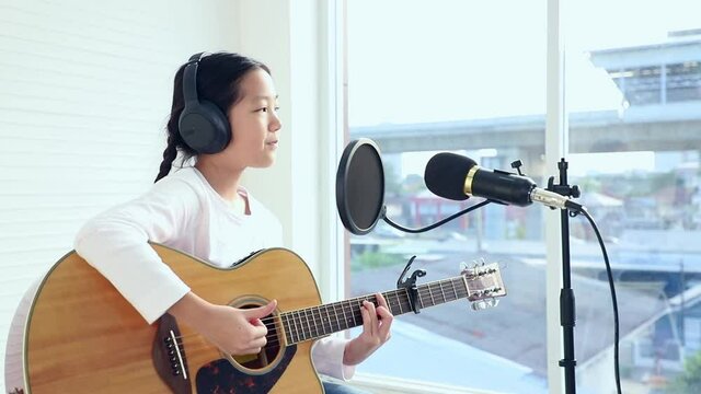 Asian young girl sitting and playing guitar in living room look over window see road and sky train station. Music lover concept.