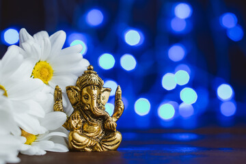 Golden lord ganesha sclupture in daisy flowers over blue illuminated background. Copy space for text