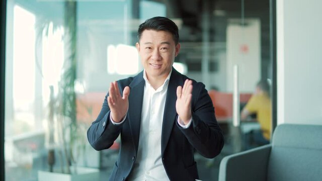 Asian Business Man Speaker In Formal Suit Talking Looking At Camera. Online Meeting, Conference Or Distance Learning, Remotely Training Via Video Call. Webcam View. Standing In A Modern Office Class