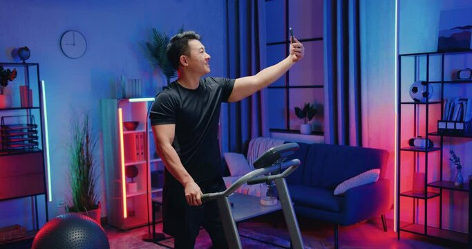 Good-looking smiling active sporty asian man in sportswear walking on treadmill and making selfie on smartphone during evening home workout,lifestyle concept