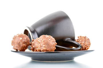 Several sweet chocolate candies with nuts on a ceramic saucer with a cup, close-up, isolated on white.