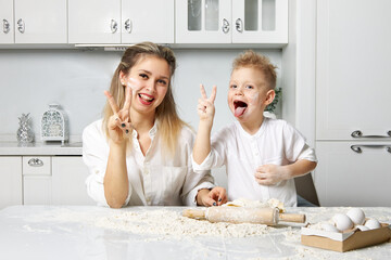 Cheerful mother and son smeared in flour in a white kitchen and show a sign of peace with their hands
