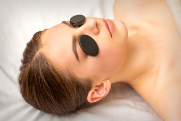 Obraz na płótnie Canvas Black massage stones lying on the eyes of the young caucasian woman. Facial massage in a spa