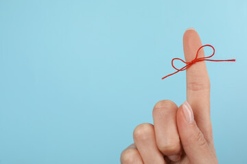 Woman showing index finger with tied red bow as reminder on light blue background, closeup. Space...