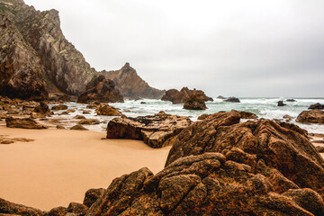 Fototapeta na wymiar Beautiful scenic landscape in cloudy weather on Ursa beach, sandy coast of the Atlantic Ocean with turquoise raging water among rocks and cliffs, Portugal