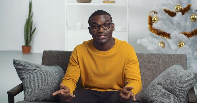 View from web camera black man Afro American businessman student wears glasses and yellow sweater sitting at home living room on couch surrounded by Christmas decoration speaks gestures talks online